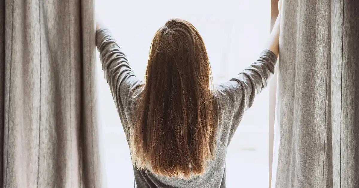 Person with long hair opening curtains, seen from behind.