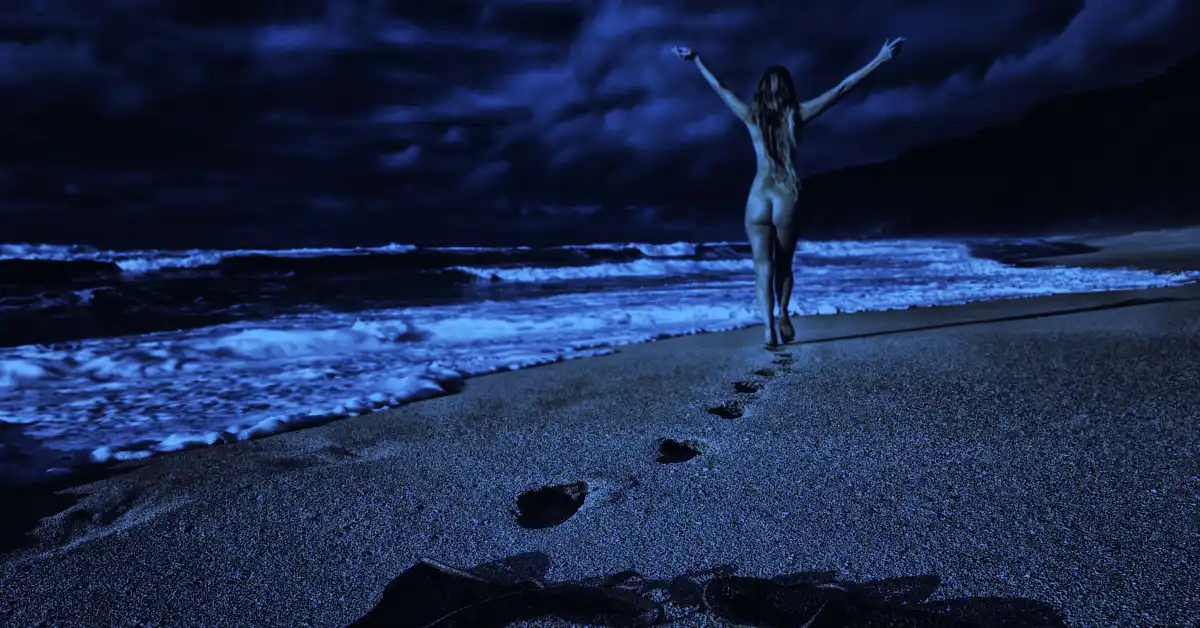 Person walking on a beach at night with arms raised, leaving footprints in the sand.