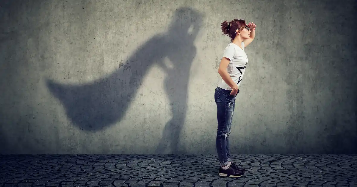 Young woman standing against a wall, with her shadow resembling a superhero with a cape.