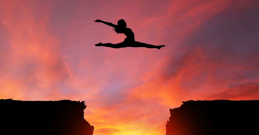 Silhouette of a person performing a mid-air split leap over a gap between two cliffs, set against a dramatic sunset sky with vibrant pink and orange hues.