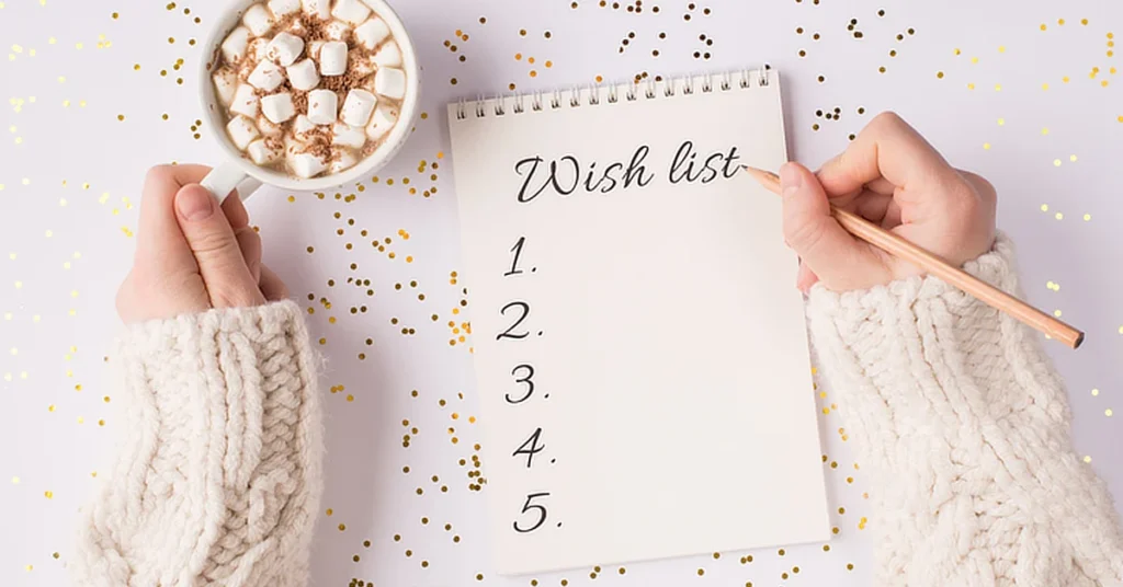 Person in a cozy sweater holding a cup of hot chocolate with marshmallows, writing a 'Wish list' in a notepad. The scene is set against a festive background with scattered gold confetti.