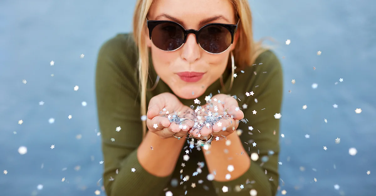 Person wearing sunglasses blowing silver star-shaped confetti from their hands, with a blurred blue background.