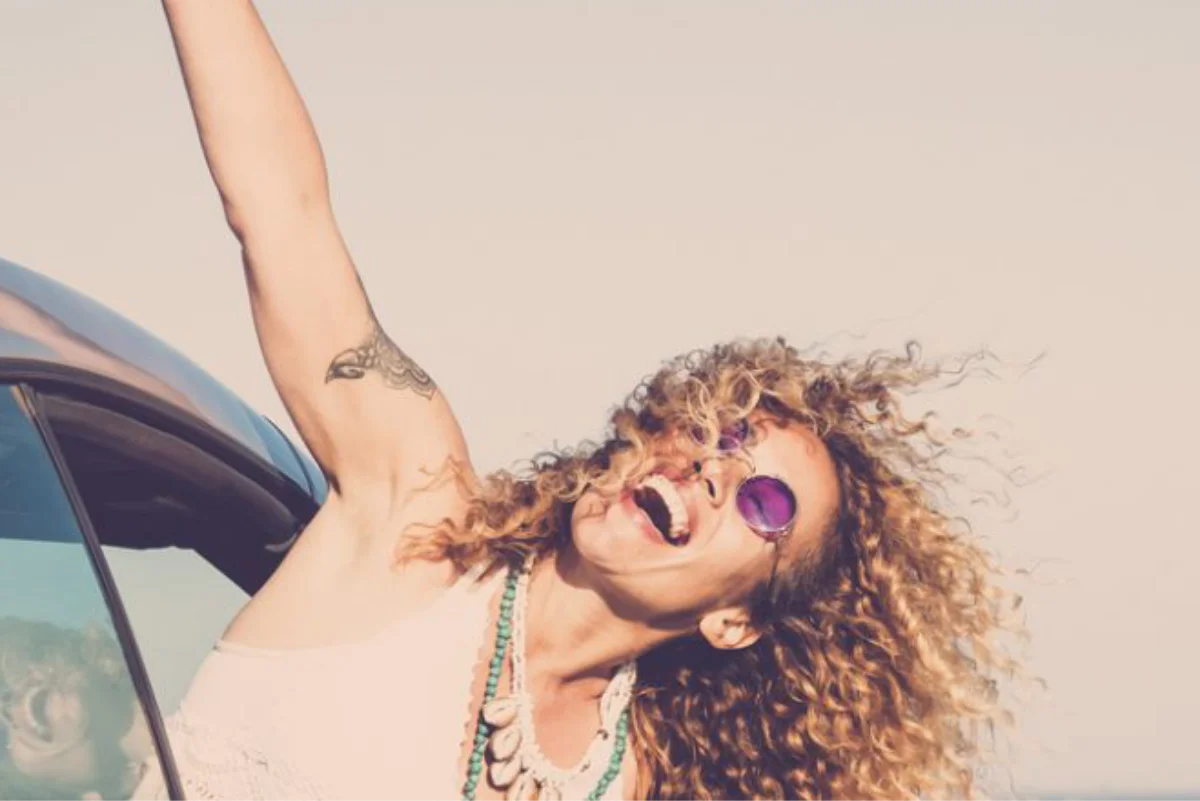 A joyful woman with curly hair and purple sunglasses leans out of a car with her arm raised, smiling against a bright sky.