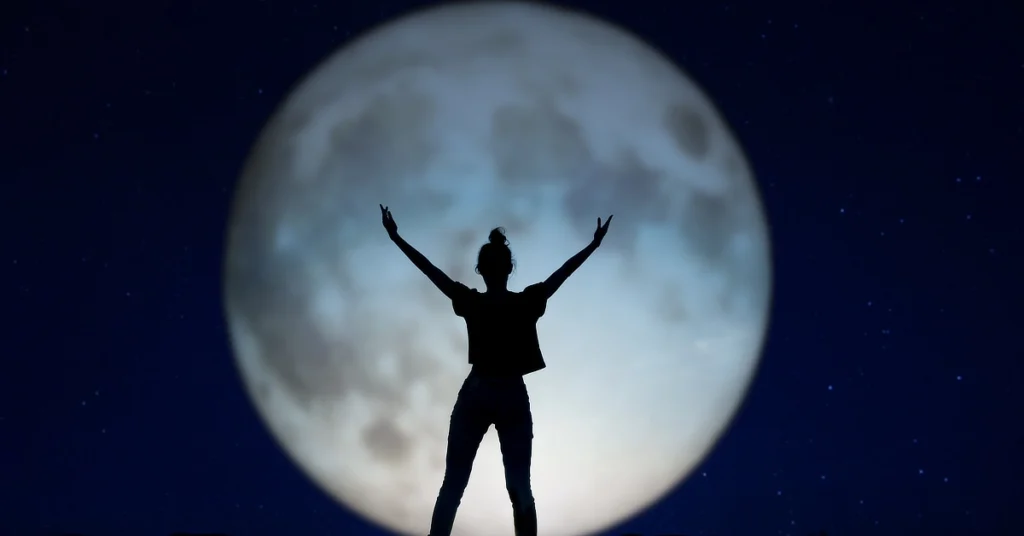 Silhouette of a person standing with arms raised, set against the backdrop of a large, full moon in a starry night sky.