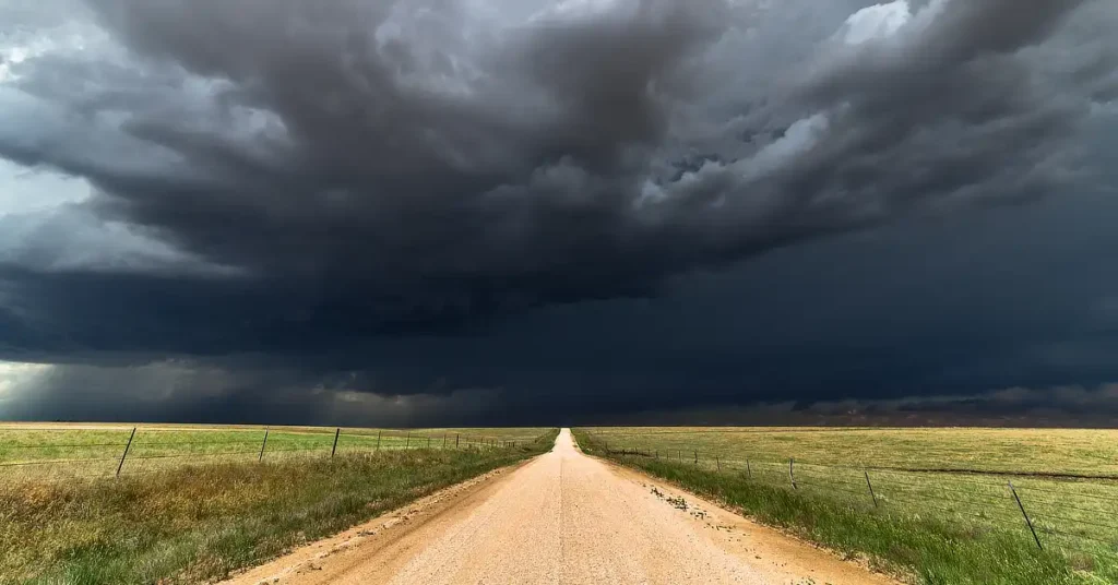 A long dirt road stretching into the distance under a sky filled with dark, ominous storm clouds, with open fields on either side.