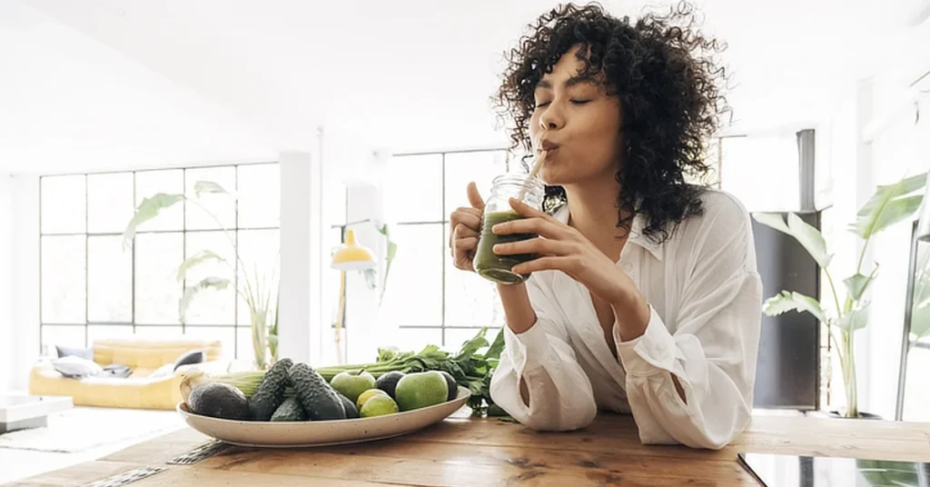 Person drinking a green smoothie at a table with a bowl of avocados and limes, in a bright, modern room with large windows and plants.