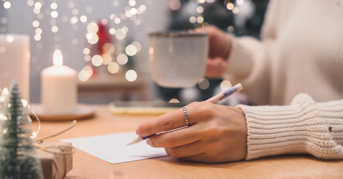 Person writing on a piece of paper while holding a mug, with a cozy, festive background featuring bokeh lights, a candle, and holiday decorations.