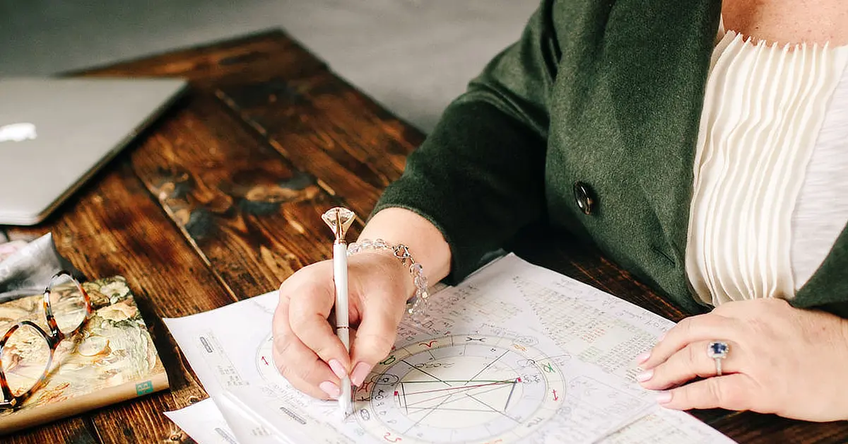 Kim Woods, Master Astrologer, seated at a wooden table, writing on a birth or natal chart with a pen. The table also holds a pair of glasses, a closed book, and a laptop.