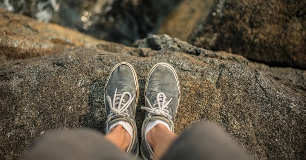 A top-down view of a person wearing worn sneakers, standing on a rocky surface near the edge of a cliff or large boulder.