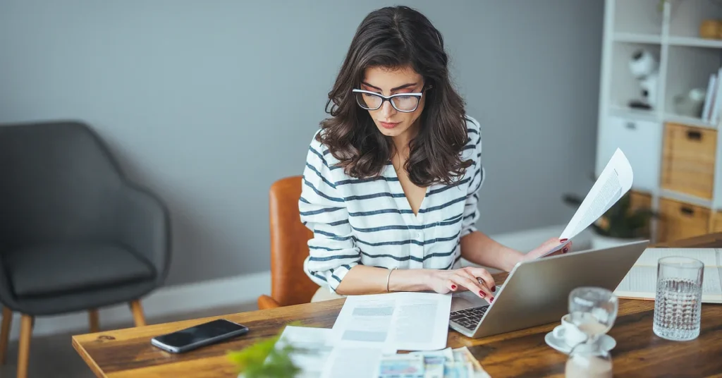 A woman with glasses and dark hair works at a desk, focused on her laptop while holding a document. The desk is covered with papers, and a smartphone is placed nearby. She is in a modern, well-lit office space.