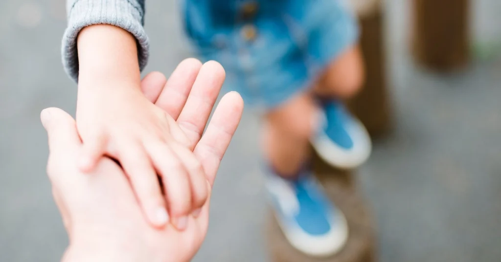 A close-up of an adult's hand gently holding a child's hand, with the child wearing blue shoes and standing in the background.