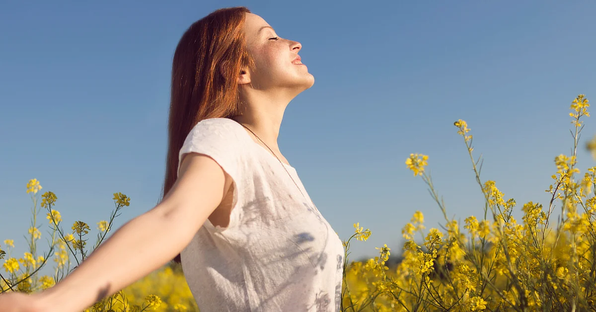 A woman with red hair stands in a field of yellow flowers with her arms outstretched, eyes closed, and face tilted up, enjoying the sunlight under a clear blue sky.