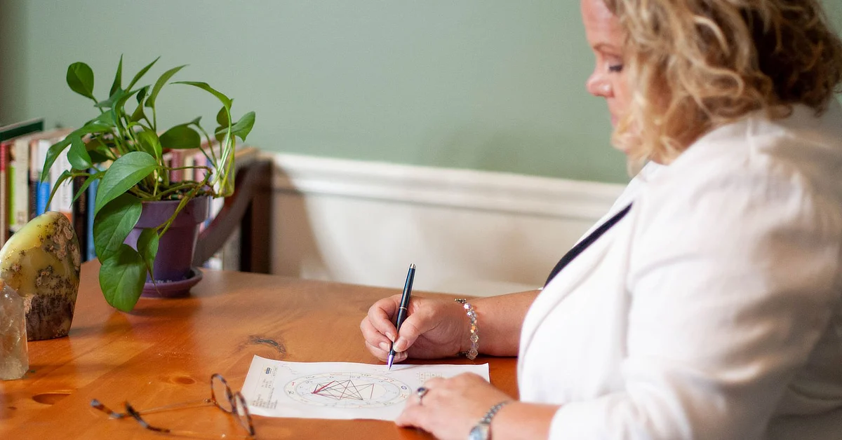 Kim Woods, Master Astrologer, seated at a wooden table, analyzing a birth or natal chart, with a pen in hand. The table also holds a potted plant, crystals, and a pair of glasses.