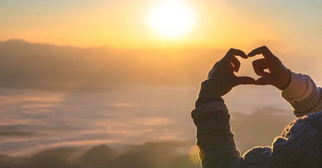 A person wearing a sweater forms a heart shape with their hands in front of a glowing sunrise, with misty mountains and a soft, cloudy sky in the background.