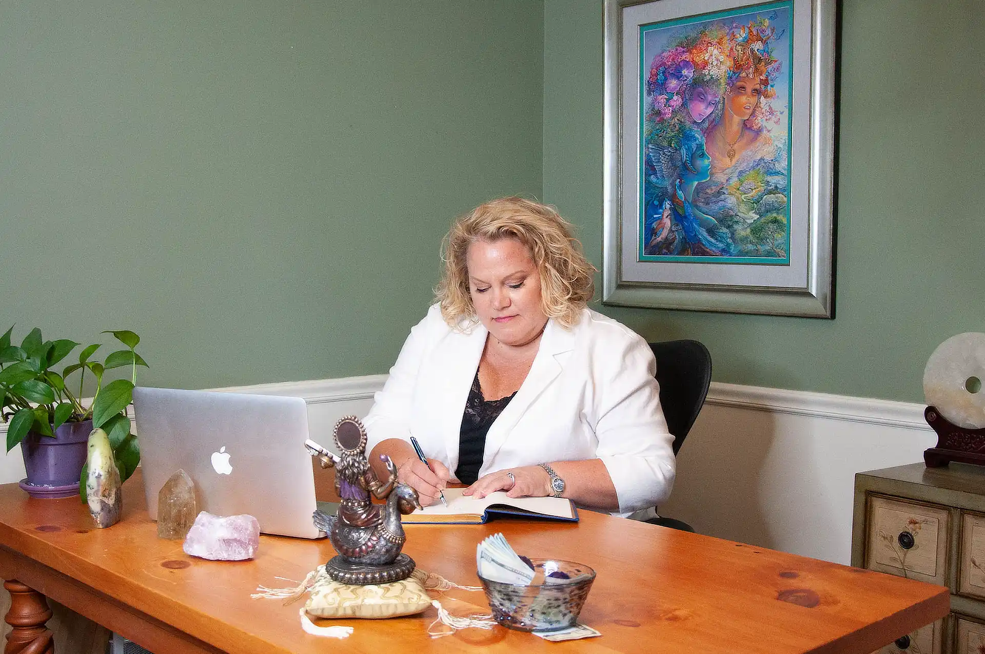 Kim Woods wearing a white blazer sits at a wooden desk, writing in a notebook. A laptop, crystals, and a small statue are on the desk. A colorful painting hangs on the green wall behind her.