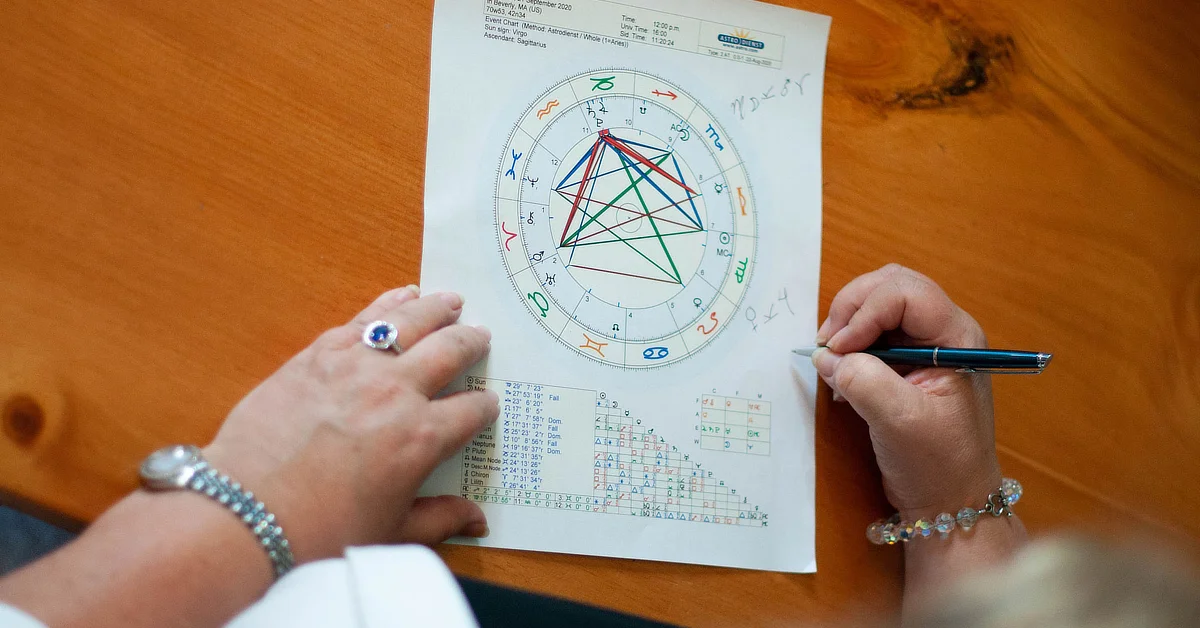 Person analyzing a printed astrological birth chart on a wooden table, holding a pen and making notes.