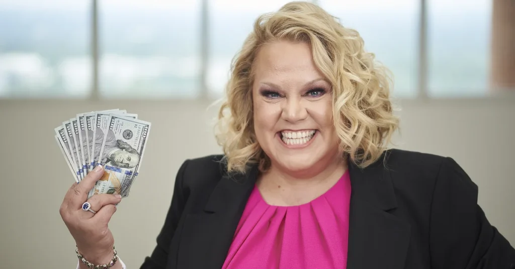 Kim Woods holds a fan of $100 bills in one hand, wearing a black blazer and a bright pink top.