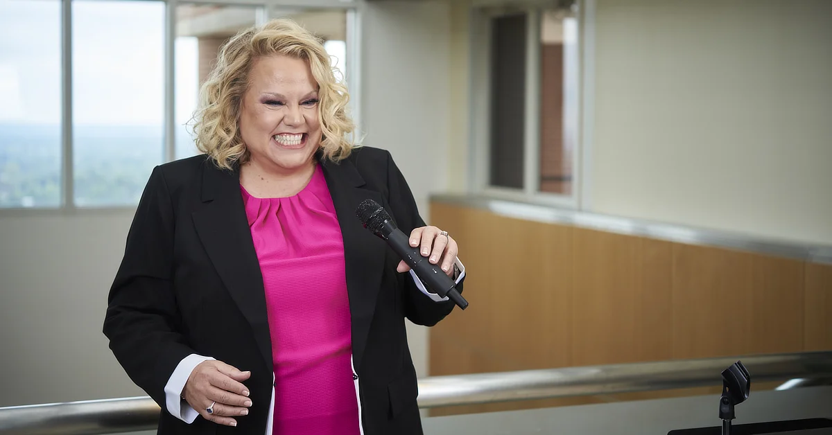 Kim Woods wearing a blazer holding a microphone and speaking to an audience indoors.