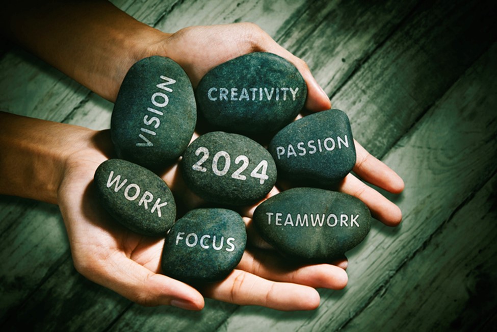 Two hands holding several smooth, black stones. Each stone has a word engraved on it, including 'VISION,' 'CREATIVITY,' 'PASSION,' 'WORK,' 'FOCUS,' 'TEAMWORK,' and '2024.' The stones symbolize motivational values and goals for the year 2024.