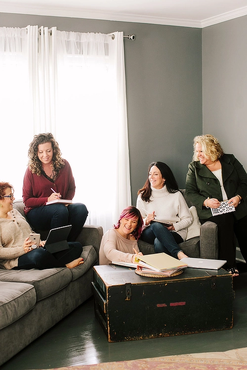 A group of five women sitting on couches and chairs in a cozy room, smiling and engaging in conversation. Some are holding notebooks, a tablet, and a laptop, suggesting a collaborative or brainstorming session.