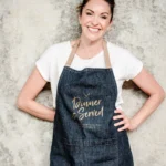 Claudia Lawrence, smiling and wearing a denim apron with 'Dinner is Served' printed on it, standing against a textured background.