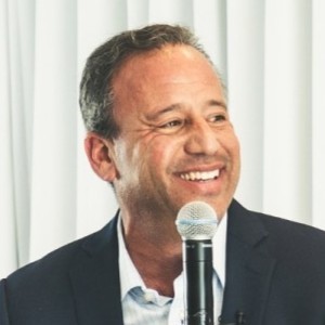 David Meltzer, smiling, with short dark hair, wearing a suit, speaking into a microphone.