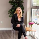 Client testimonial image featuring HIllary Gadsby, a woman, in a black suit sitting on a clear chair, smiling confidently in a modern office setting with a vase of pink roses nearby.