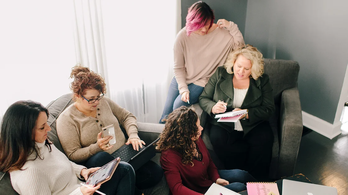 Kim Woods and a group of clients gathered in a cozy setting during an in-person retreat. They are engaged in discussion, taking notes, and using laptops and tablets.