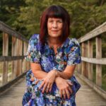 "Client testimonial image of Maura Allard in a colorful floral dress sitting on a wooden bridge, surrounded by lush greenery.