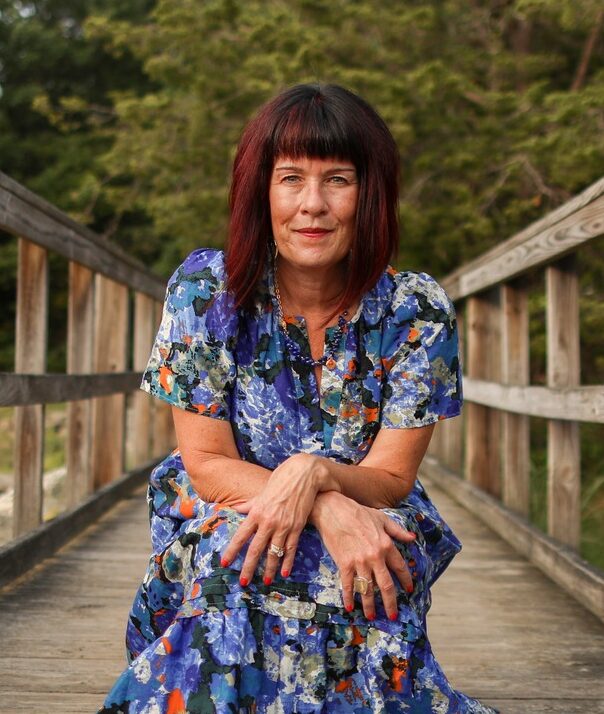 "Client testimonial image of Maura Allard in a colorful floral dress sitting on a wooden bridge, surrounded by lush greenery.