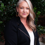 Client testimonial image featuring a woman, Miriam Chadwell, with long blonde hair, wearing a black jacket and white v-neck blouse, smiling outdoors with trees in the background.