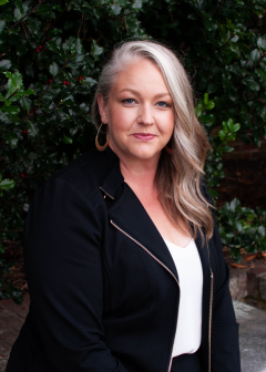 Client testimonial image featuring a woman, Miriam Chadwell, with long blonde hair, wearing a black jacket and white v-neck blouse, smiling outdoors with trees in the background.