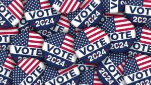 A collection of round "Vote 2024" buttons featuring the American flag design. The buttons have the word "VOTE" in bold white letters and "2024" below, with a red check mark inside the "0" in "2024." The buttons are stacked and scattered, creating a visually layered pattern