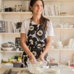 Client testimonial image featuring Paula Montenegro in a patterned apron mixing dough in a glass bowl, standing in a warmly lit kitchen with shelves of books and dishware in the background.