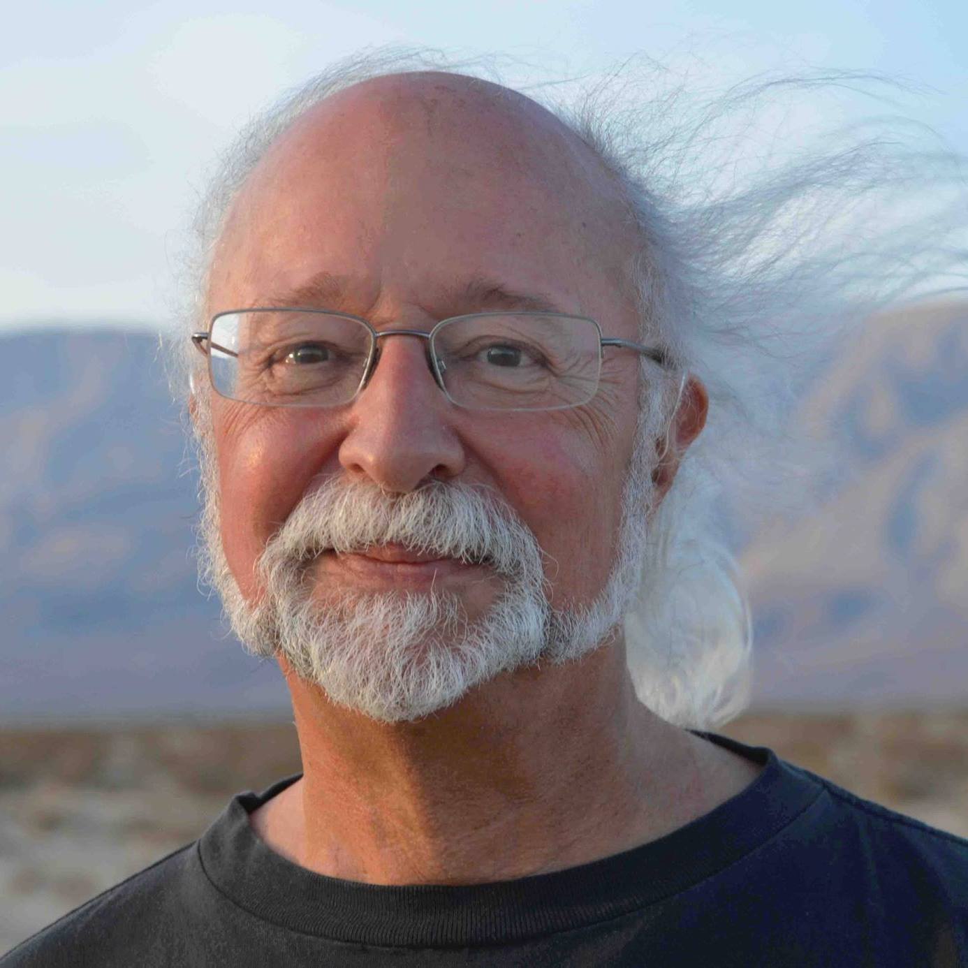 Steven Forrest, with long white hair, a beard, and glasses, standing outdoors with a desert landscape in the background.