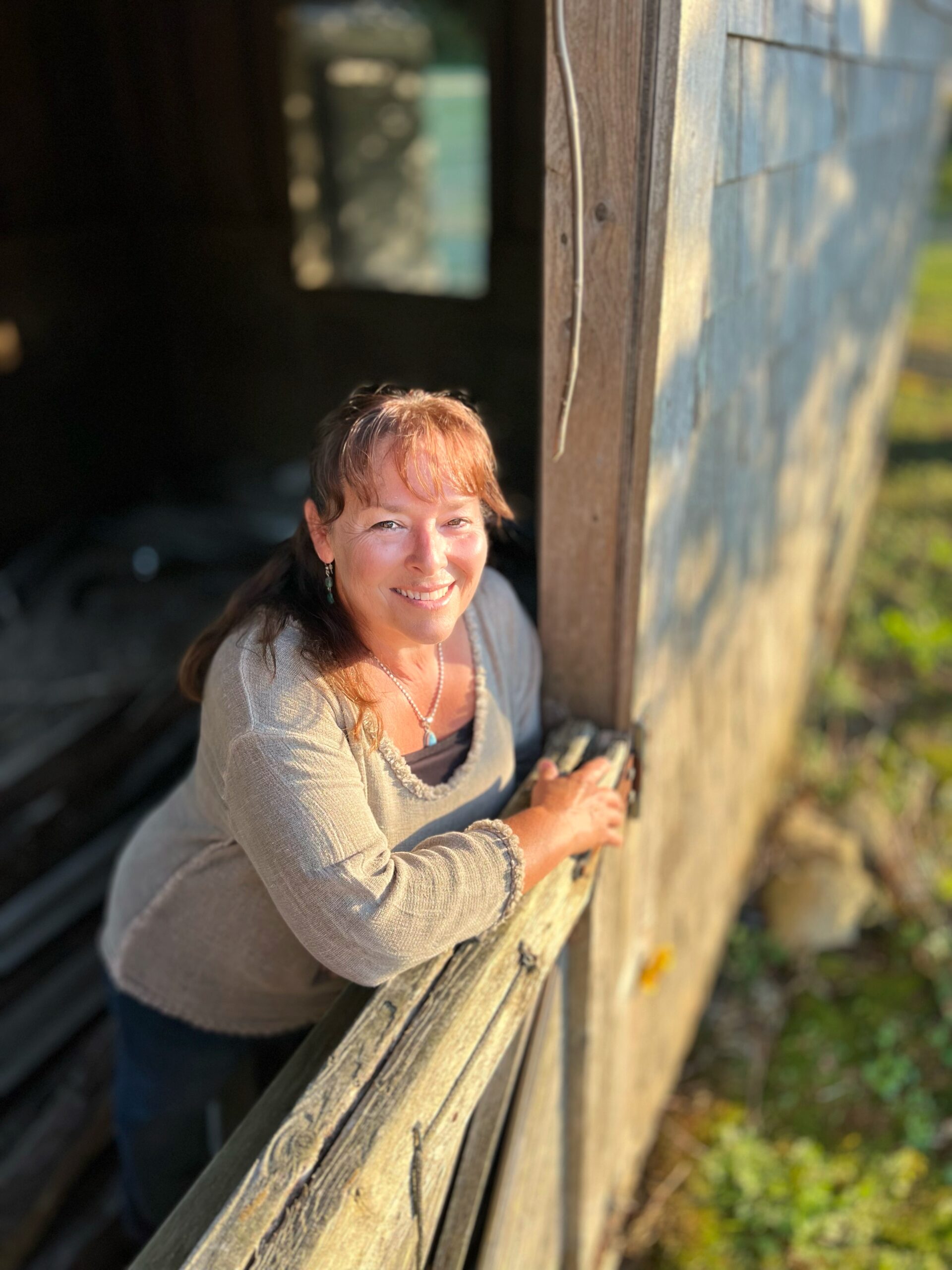 leans on a rustic wooden fence, smiling warmly in the sunlight, with a weathered wooden building and greenery in the background.