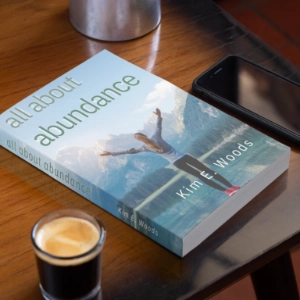 "All About Abundance" by Kim E. Woods sitting on a wooden table next to a cup of coffee and a smartphone.