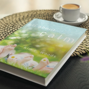 A hardcover copy of "All About Calm" by Kim E. Woods resting on a woven mat beside a cup of coffee.