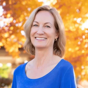 Amy Collette wearing a blue top, standing outdoors in front of vibrant autumn foliage.