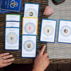 A spread of Intuition Oracle Cards laid out on a wicker table, with a hand pointing to one of the cards and a rose quartz crystal nearby.