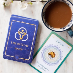 A deck of Intuition Oracle Cards on a marble surface, with one card ("Fortune") displayed next to a cup of coffee in a floral-patterned mug.