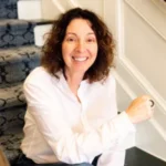 Iris Goldfeder wearing a white shirt sits on a carpeted staircase, smiling at the camera.