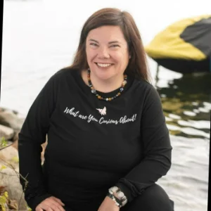 Kelly Wagner smiling while sitting by the water, wearing a black top that reads 'What are you curious about?' and a beaded necklace.