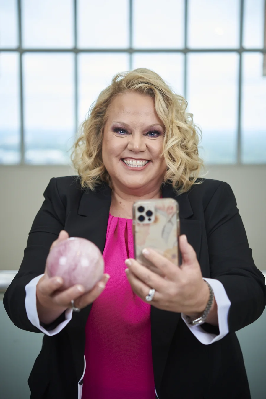 Kim Woods wearing a black blazer and pink blouse, holding a smartphone in one hand and a pink crystal sphere in the other, standing in front of a large window.