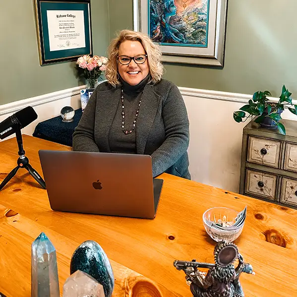 Kim Woods smiles while sitting at a wooden table with a laptop in front of her. She is wearing a gray turtleneck sweater and a beaded necklace. A microphone is positioned on the table, along with various crystals and decorative items. Behind her, there is a framed certificate on the wall, a framed artwork, and a small cabinet with plants.