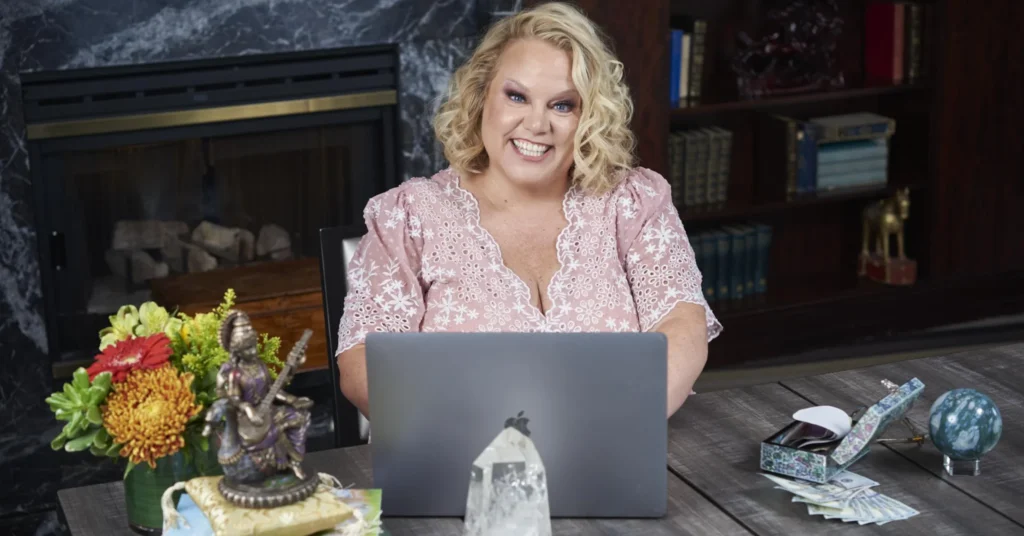 Kim Woods sitting at a table with a laptop, smiling. The setting includes a cozy fireplace, bookshelves, and spiritual elements like a crystal, a statue, and tarot cards.