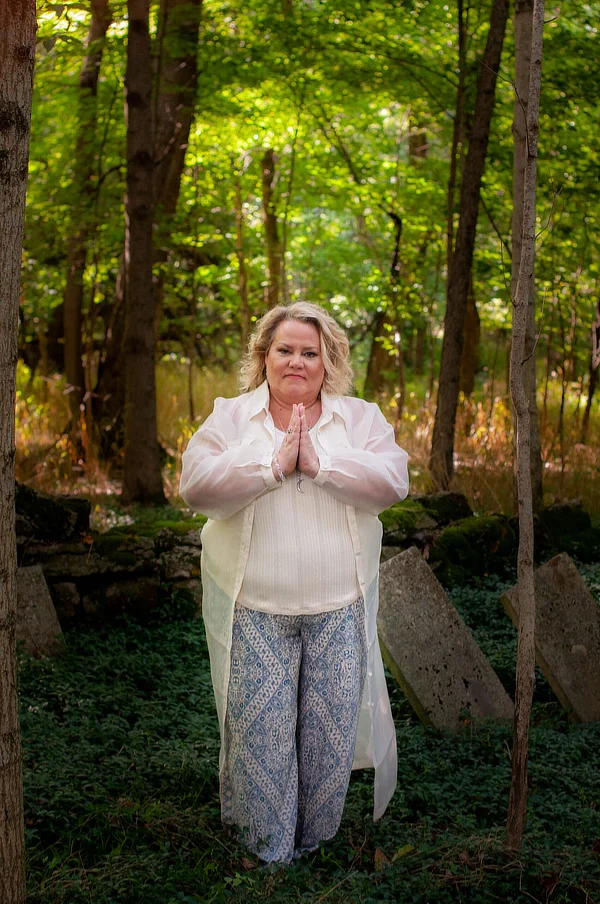 Kim Woods stands in a forest with her hands in a prayer position, wearing a white shirt and patterned pants. Sunlight filters through the trees, casting a soft glow on the greenery around her.