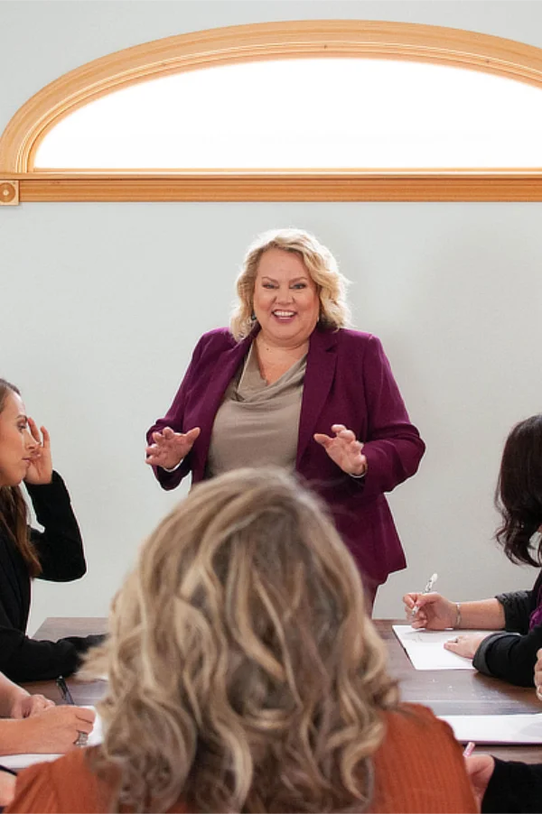 Kim Woods wearing a purple blazer speaks to a group of people seated around a table, leading a discussion or presentation.