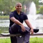 Client testimonial image featuring Matt Dillingham, a man in a black polo shirt and dark pants, sitting on a bench by a lake with a fountain in the background.