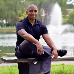 Client testimonial image featuring Matt Dillingham, a man in a black polo shirt and dark pants, sitting on a bench by a lake with a fountain in the background.